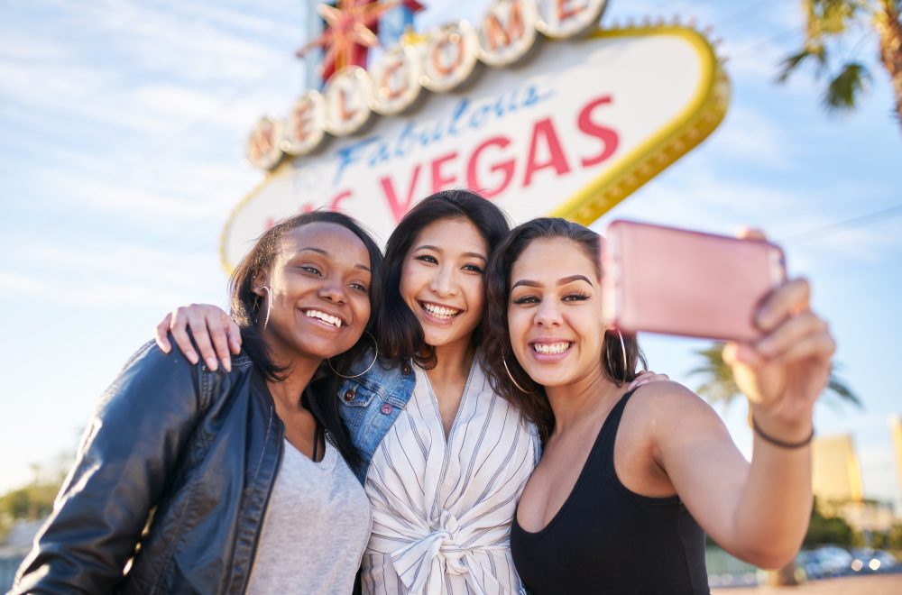 three friends on vacation taking a group selfie in front of the welcome to las vegas sign